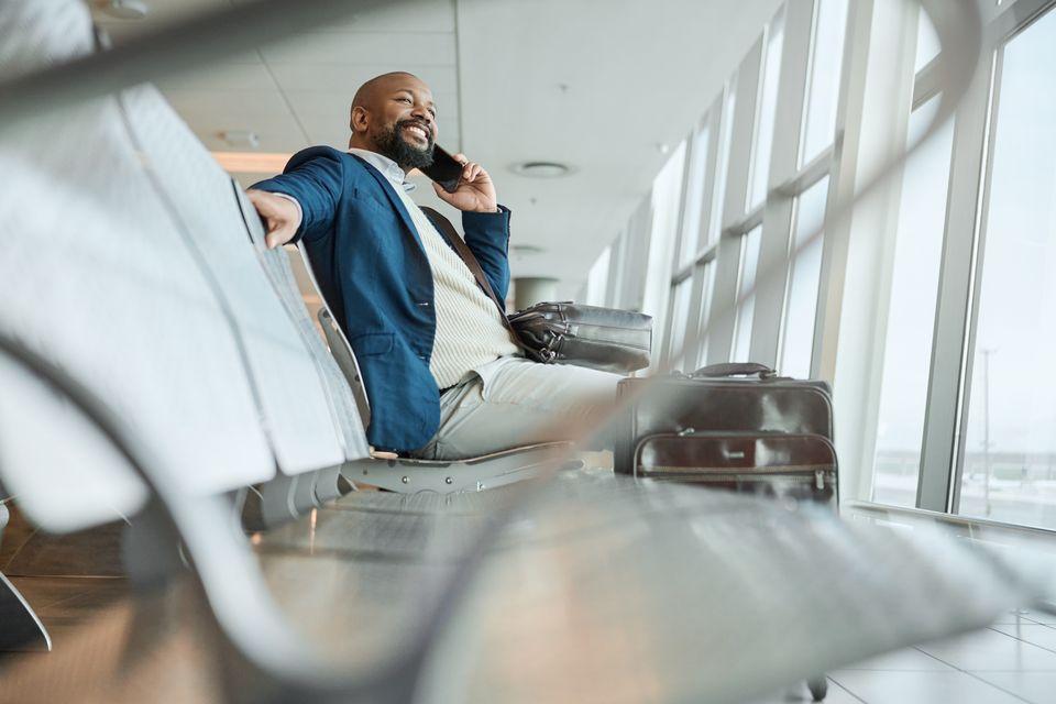 Mann der am Flughafen wartet und auf einer Bank sitzt und dabei telefoniert