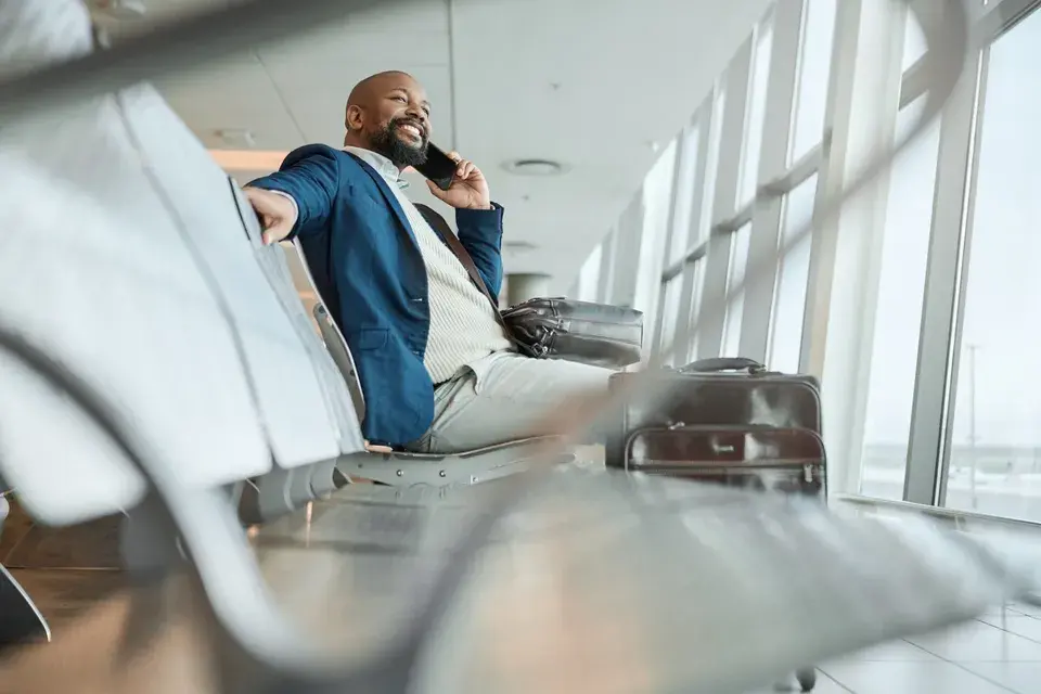 Mann der am Flughafen wartet und auf einer Bank sitzt und dabei telefoniert